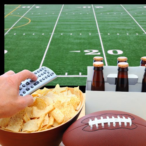 Closeup Of A Man's Hand Holding A TV Remote With A Bowl Of Chips And A Six Pack Of Beer With A Football Field On The Television Screen In The Background. Square Format.