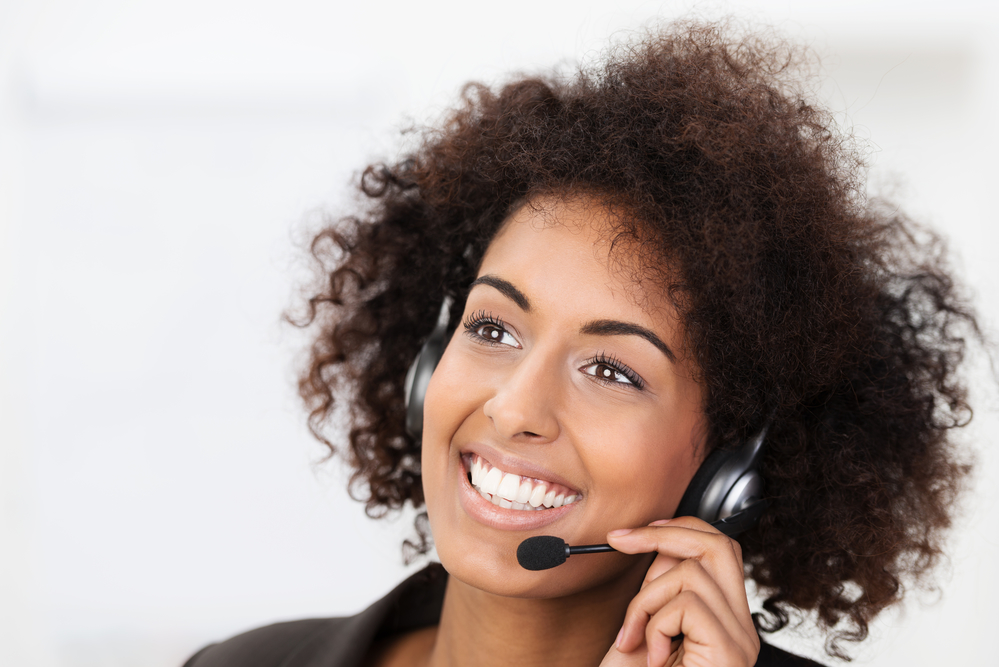 Beautiful Vivacious Young African American Client Services, Call Centre Operator Or Receptionist Smiling A Warm Friendly Natural Smile As She Listens To A Client Speaking On Her Headset